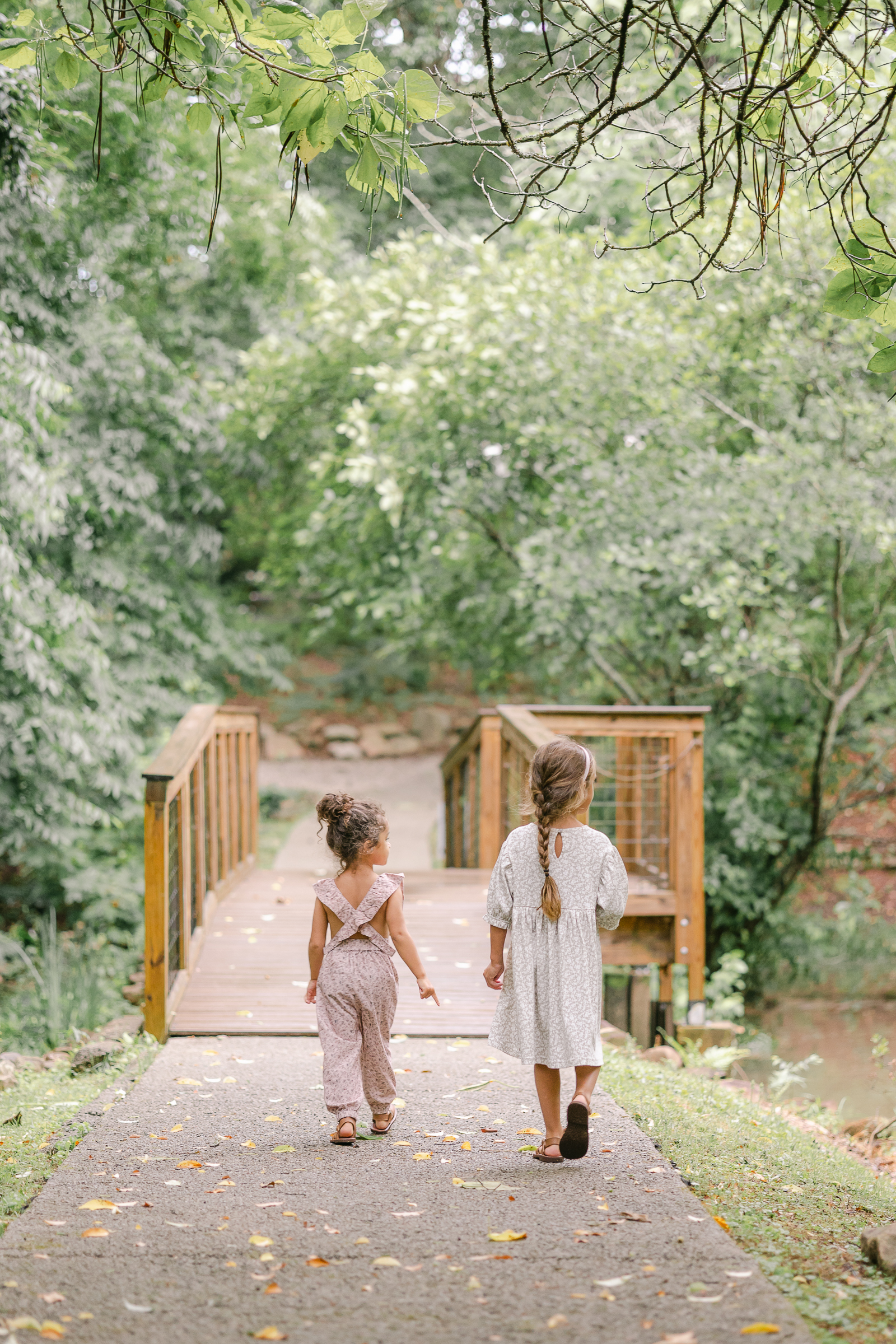 two girls walking onto a bridge surrounded by lush trees and a small pond