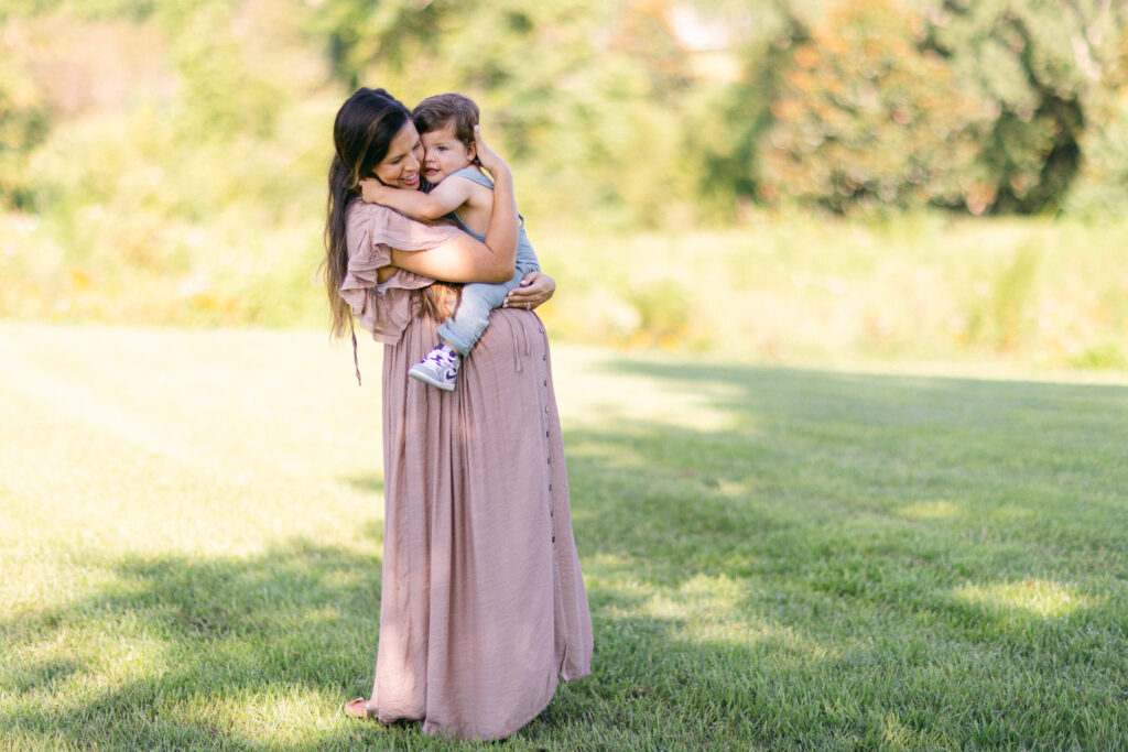 A pregnant mother in a flowing feminine dress embraces a chubby toddler on her belly in a patch of shade at Ag Heritage Park at Auburn University. 